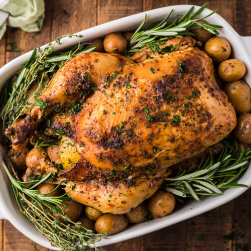 Whole beer chicken on a platter with rosemary and potatoes.