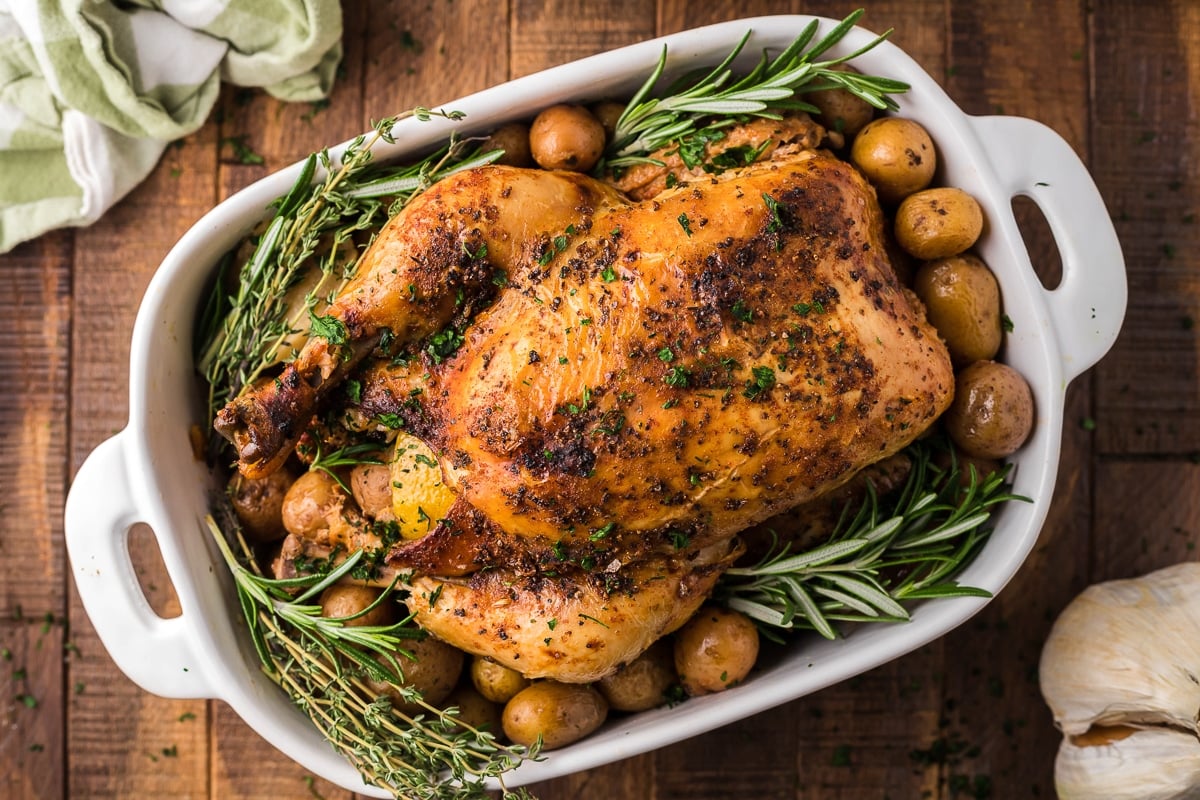 Whole beer chicken on a platter with rosemary and potatoes.