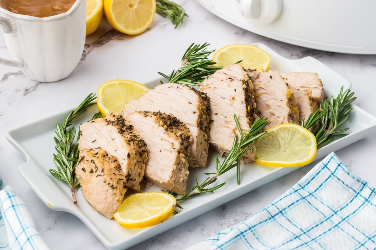 A white rectangular dish displays sliced, seasoned roast turkey garnished with fresh rosemary and lemon slices. The dish is set on a marble countertop with a blue and white checked cloth napkin nearby. A cup of gravy and whole lemons are in the background.