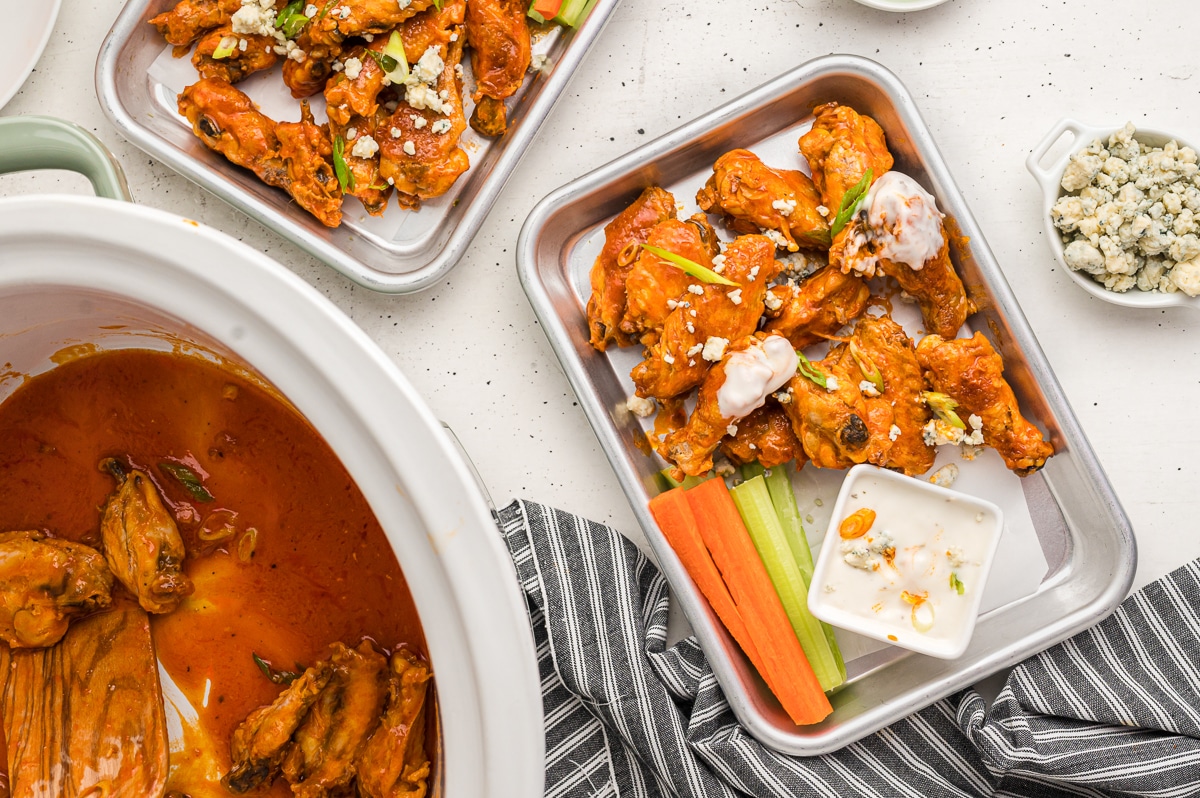 overhead shot of buffalo wings in slow cooker and on metal serving trays.