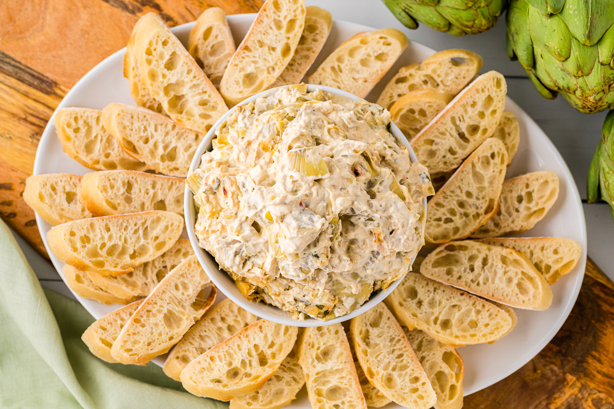 artichoke dip in a bowl with bread on a platter.