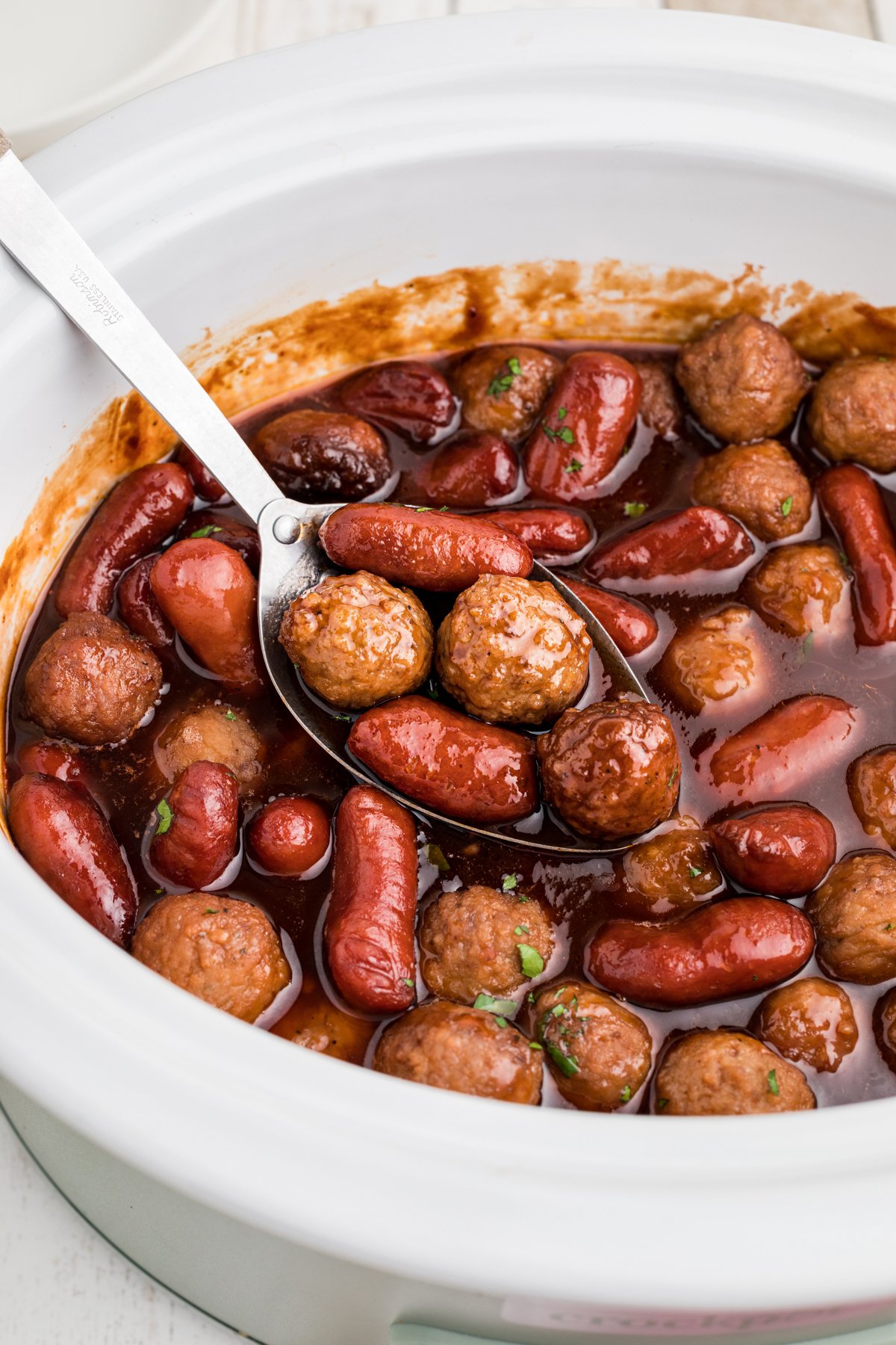 close up of meatballs and little smokies in a white slow cooker.