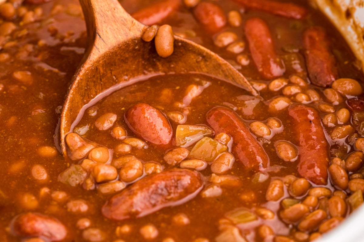 close up of baked beans and little smokies on a wooden spoon.