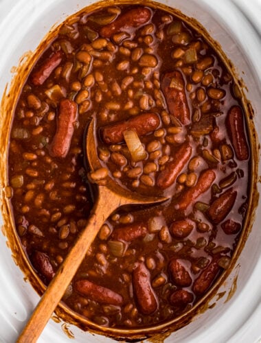 Baked beans and little smokies in a slow cooker with a wooden spoon.