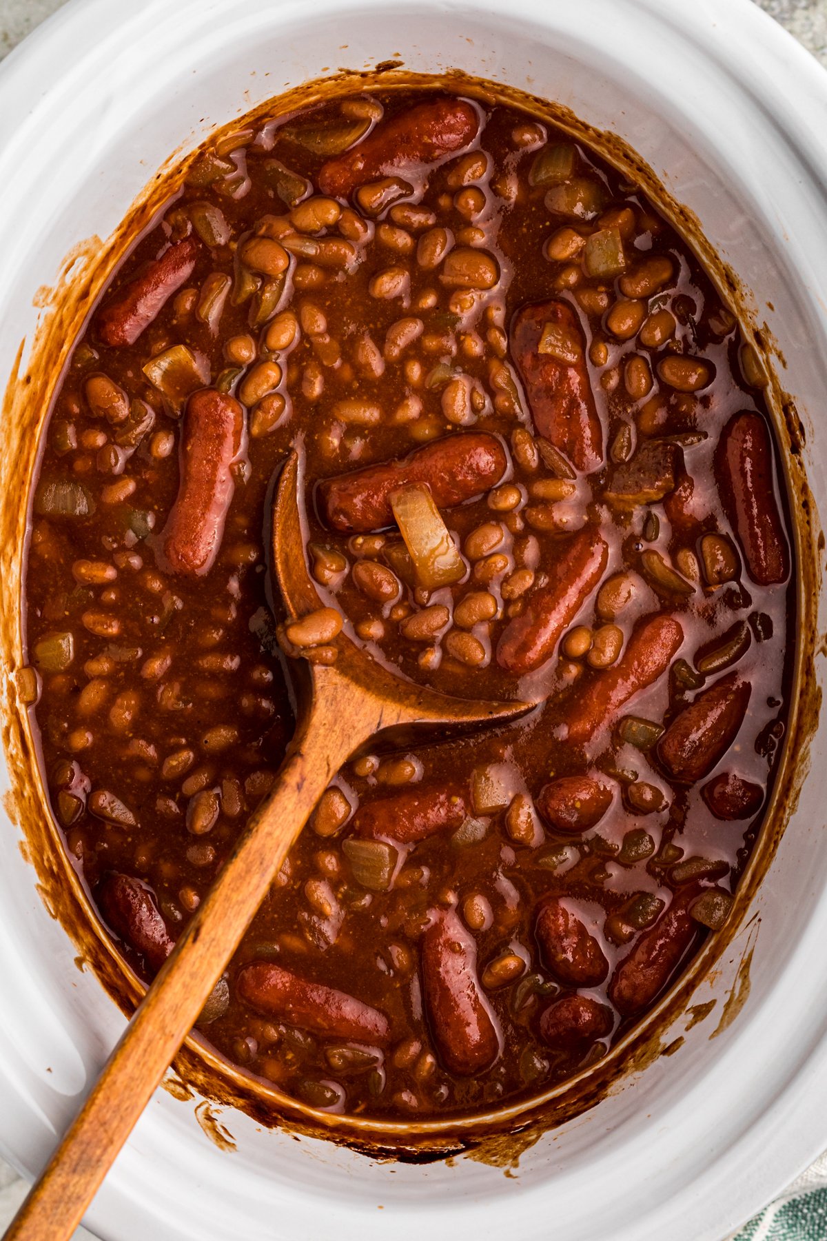 Baked beans and little smokies in a slow cooker with a wooden spoon.