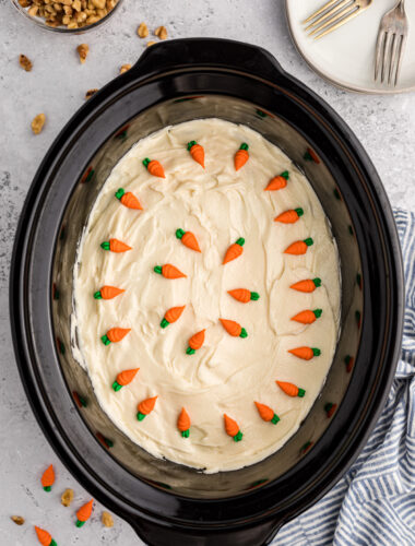 carrot cake with icing and carrots on top in a slow cooker.