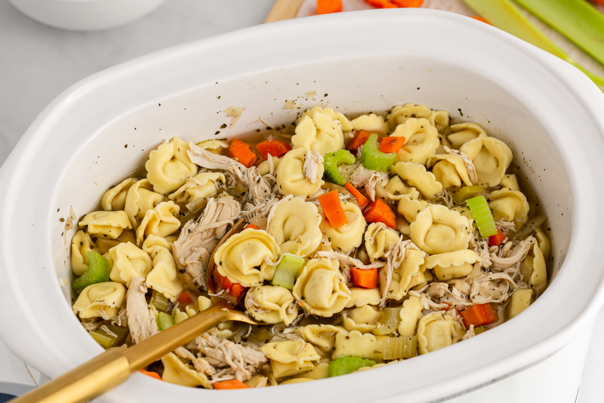 chicken tortellini in a white slow cooker with a wooden spoon in it.