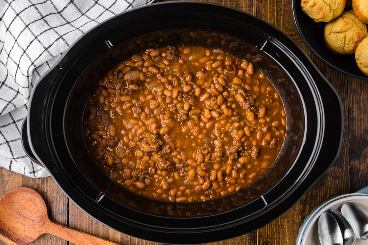 cooked cowboy baked beans in a crockpot with corn muffins on the side.