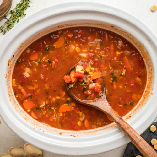 Red clam chowder in a white slow cooker with a wooden spoon in it.