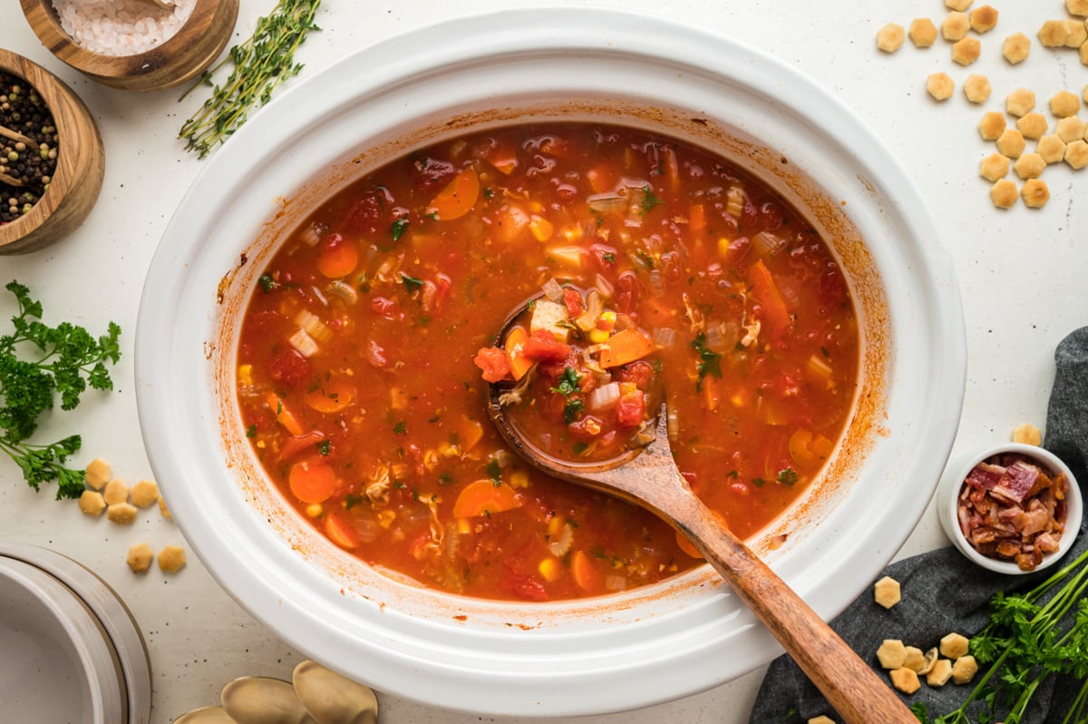 Red clam chowder in a white slow cooker with a wooden spoon in it.