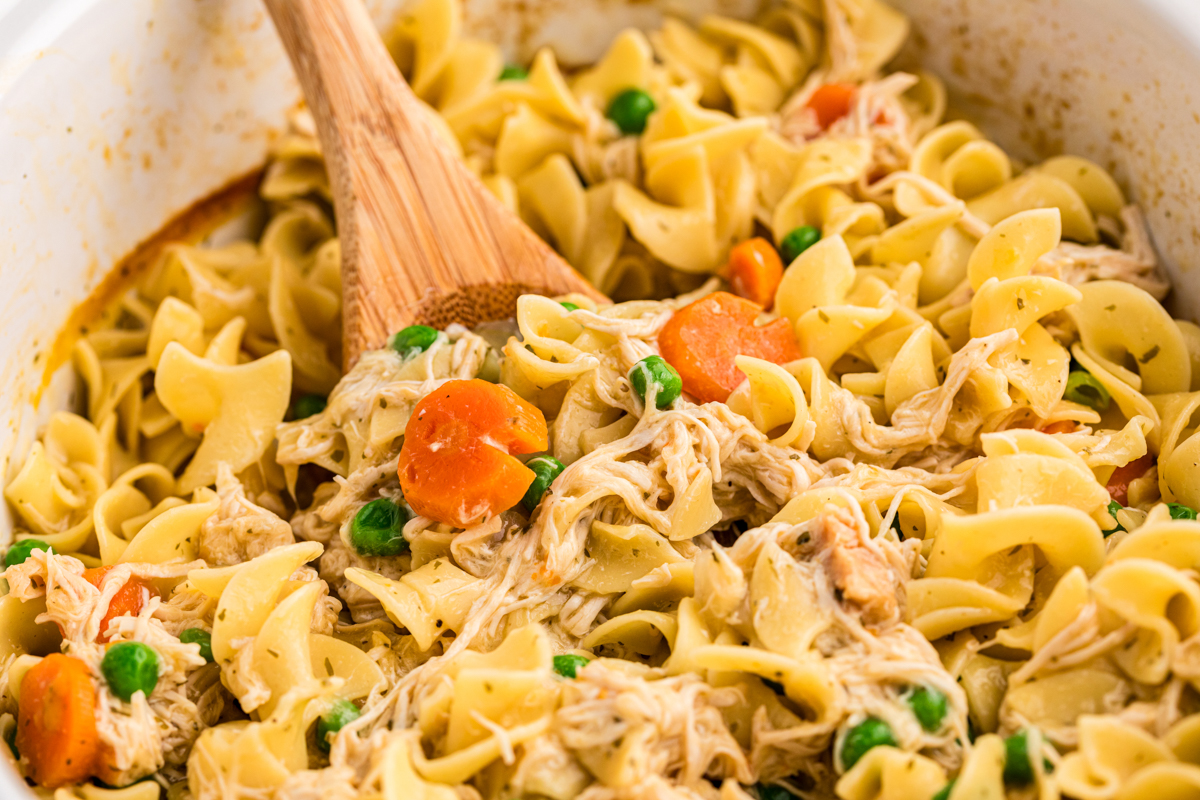 Close up of chicken and noodles in a slow cooker with a wooden spoon in it.