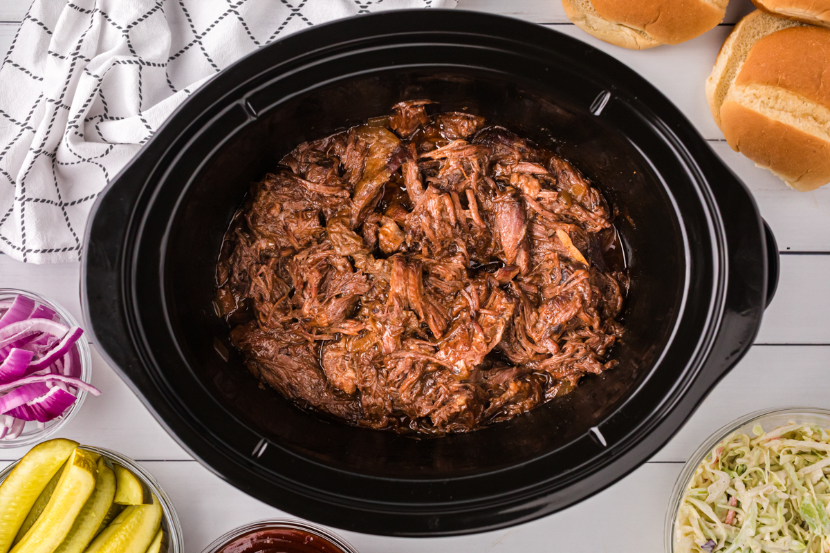 shredded bbq beef in a crockpot with pickles and slaw on the side.