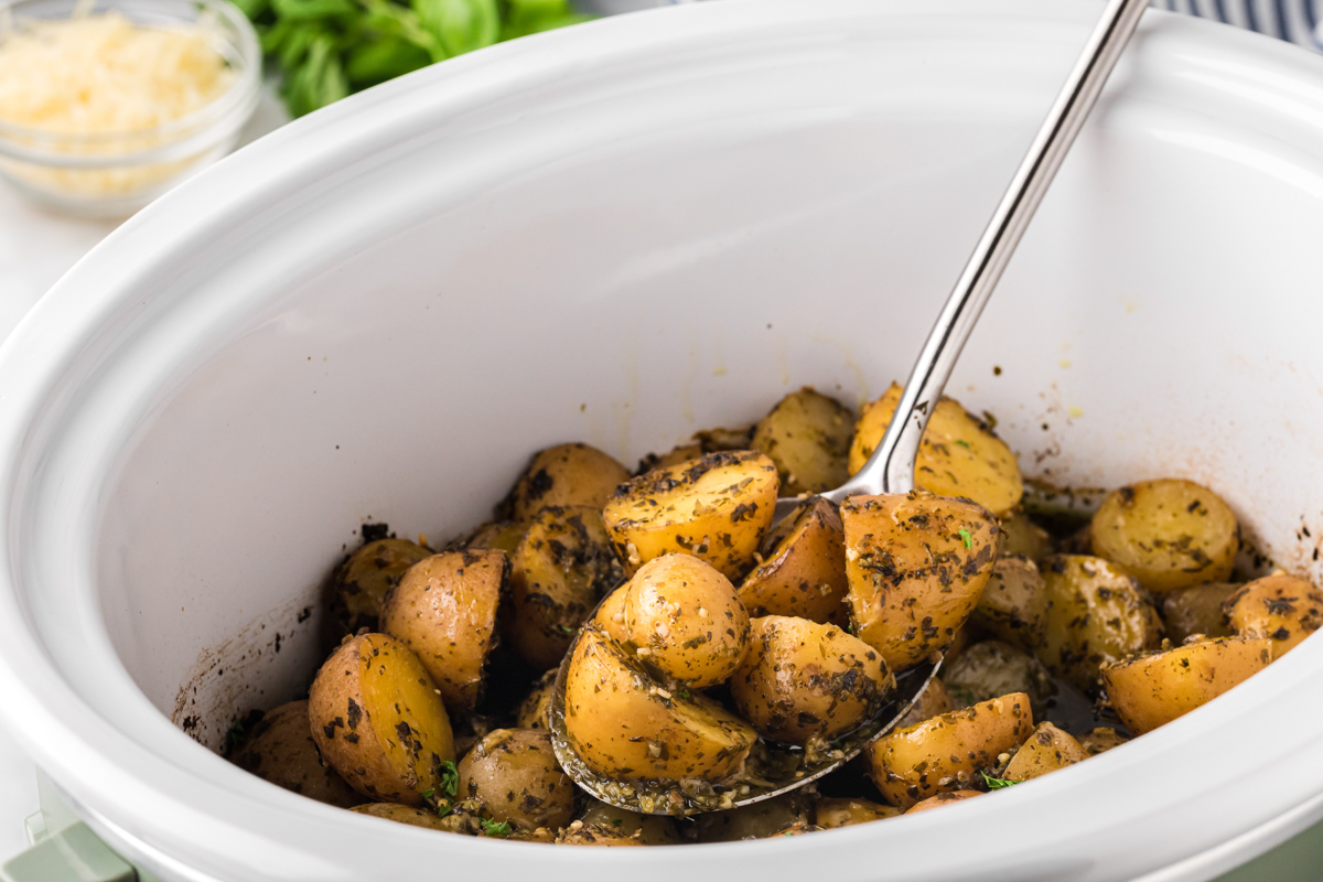 Pesto parmesan potatoes in a white slow cooke with spoon in it.