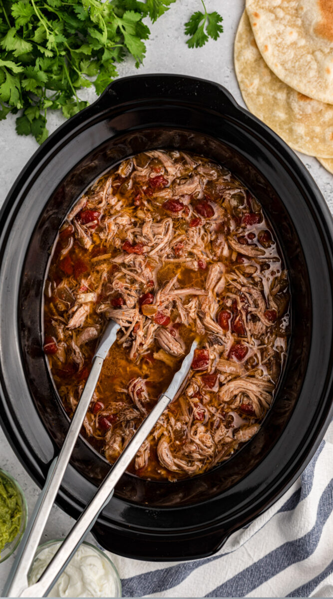 Long image of shredded pork in a slow cooker with tongs.