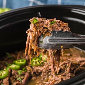 tongs pulling out some of the cooked meat from the crockpot.