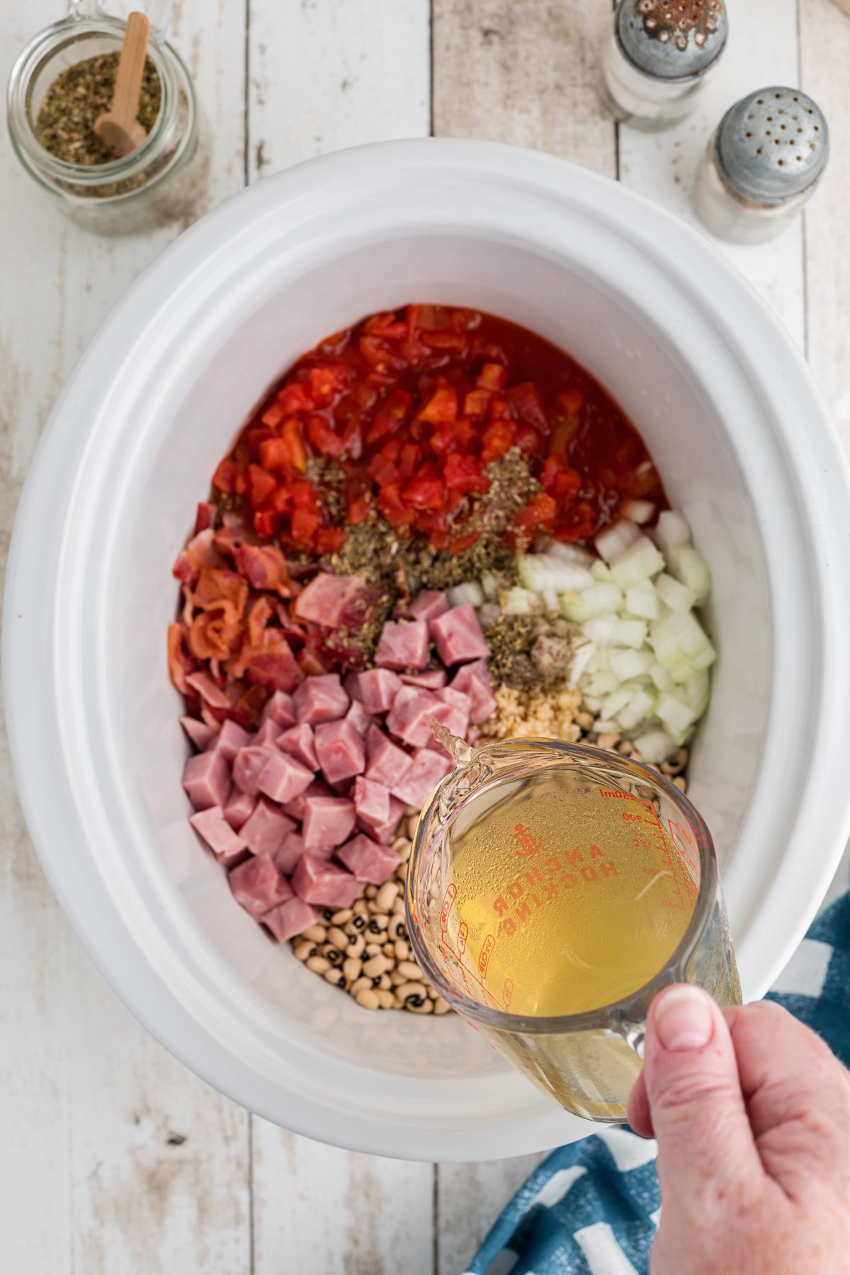 black eyed peas, vegeatbles, tomatoes, ham and bacon in slow cooker with broth being poured over.