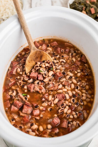 Black eyed peas in a white slow cooker with a wooden spoon in it.