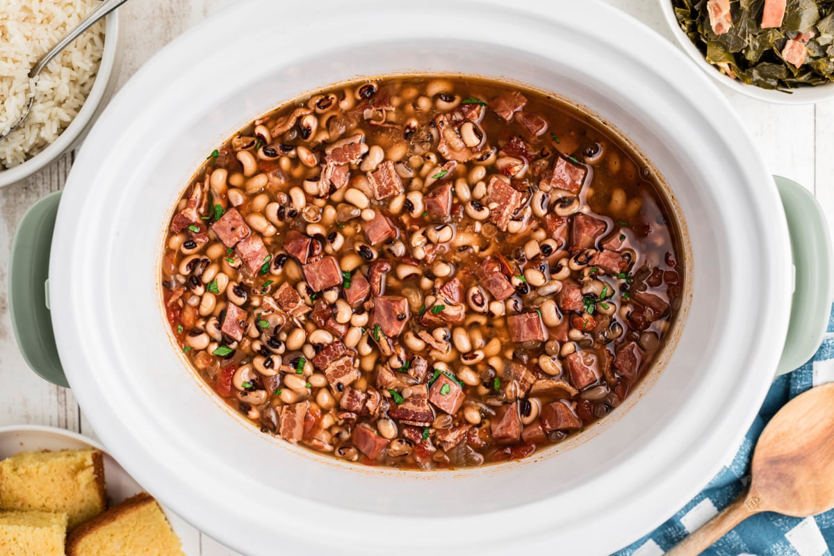 Cooked black eyed peas with ham in a slow cooker with collards and rice next to them.