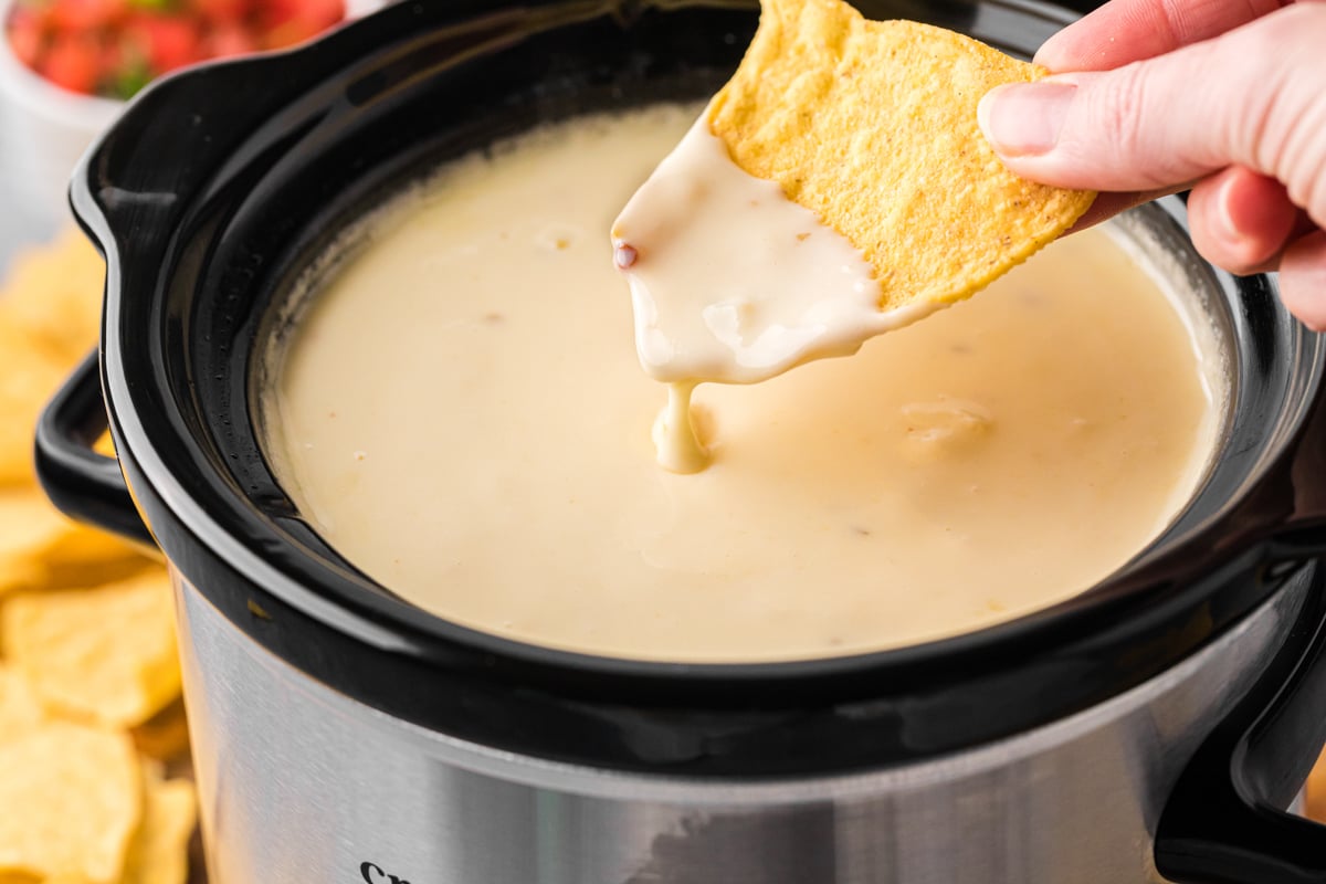 dipping a tortilla chip into restaurant style queso in a slow cooker.