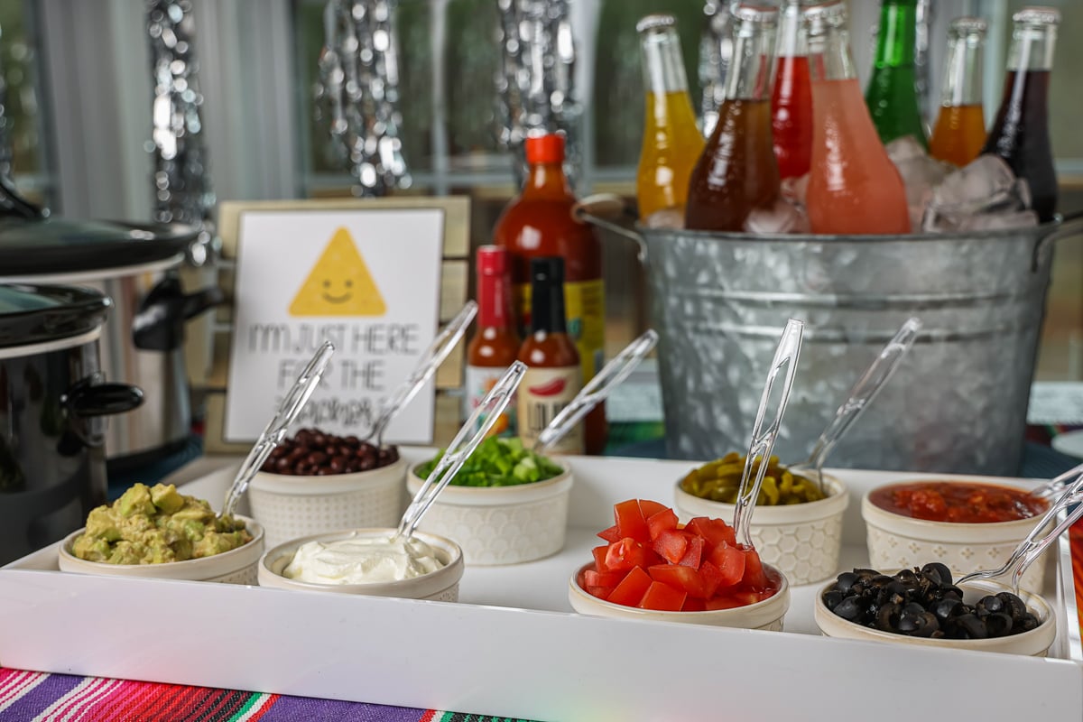 Ingredients for a nacho bar with drinks in the background.