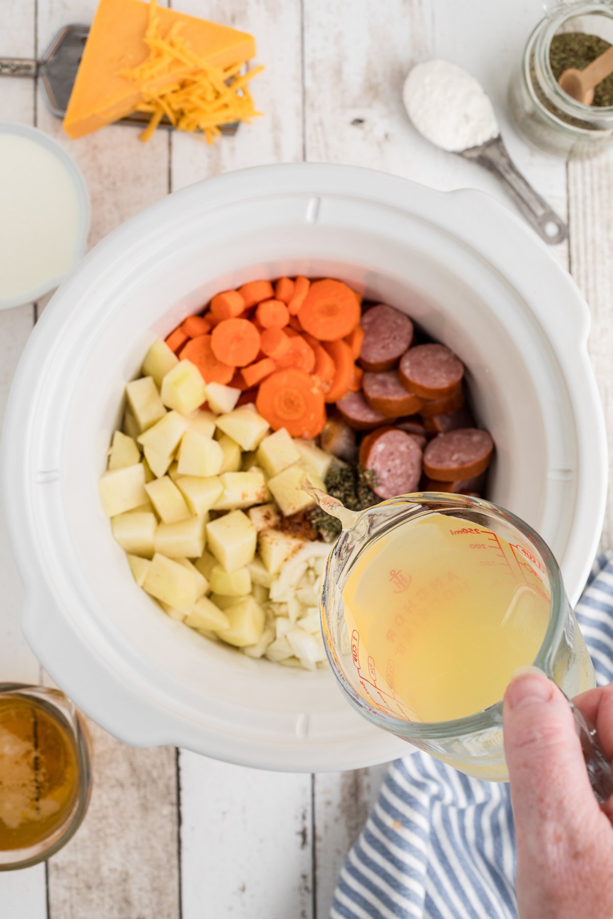 chicken broth being pour over veggies and kielbasa in a slow cooker.