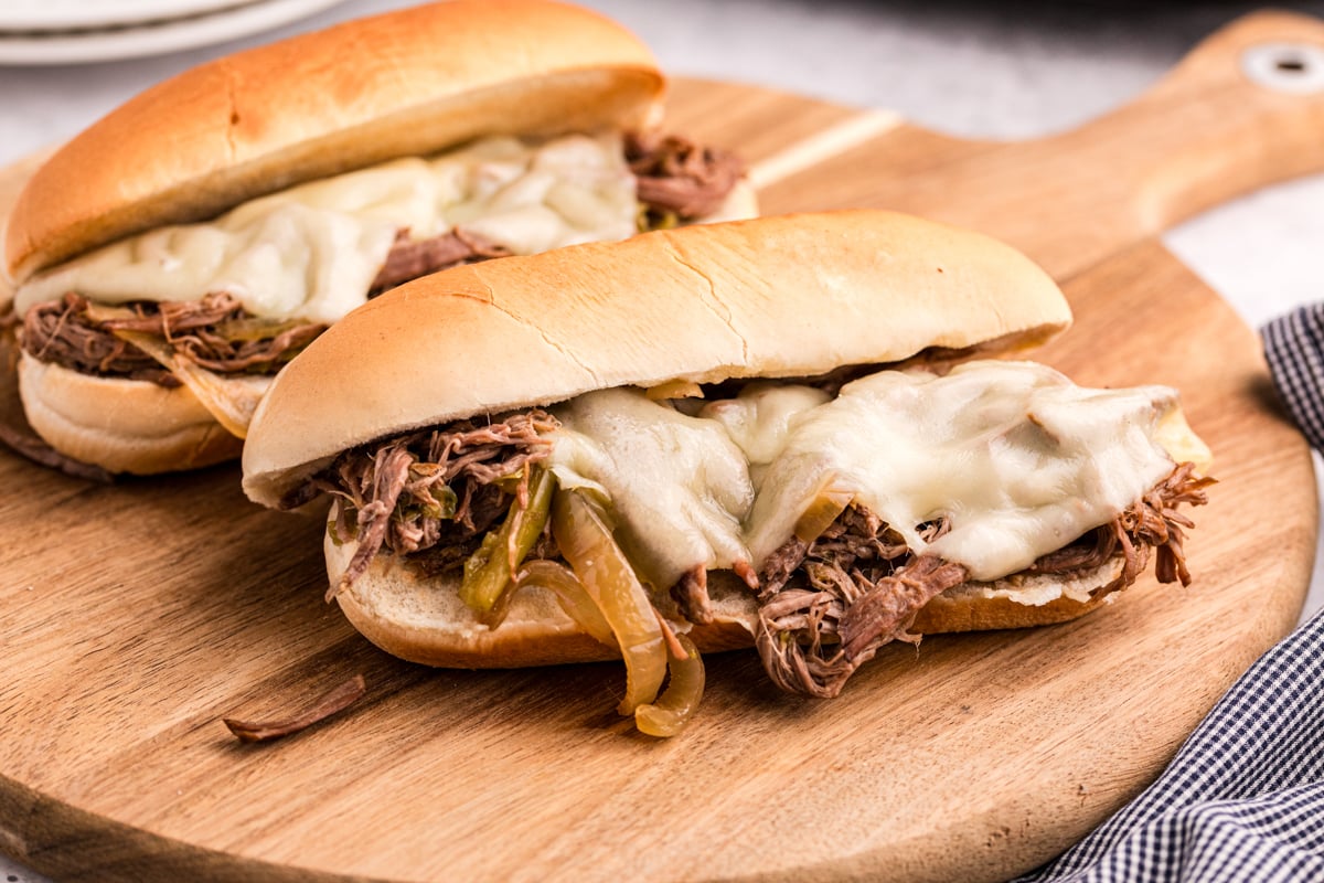 two slow cooker philly cheese steak sandwiches on a wooden board.