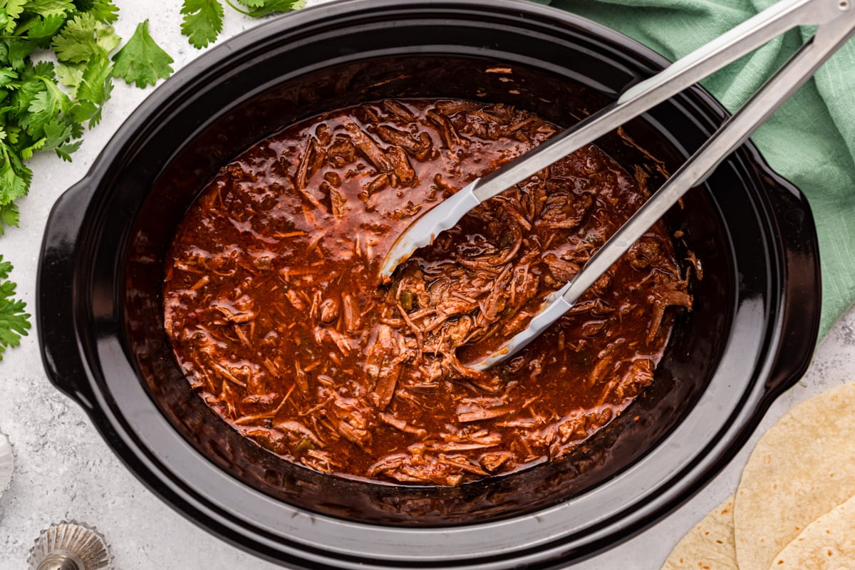 Salsa beef in a slow cooker with tongs in it.