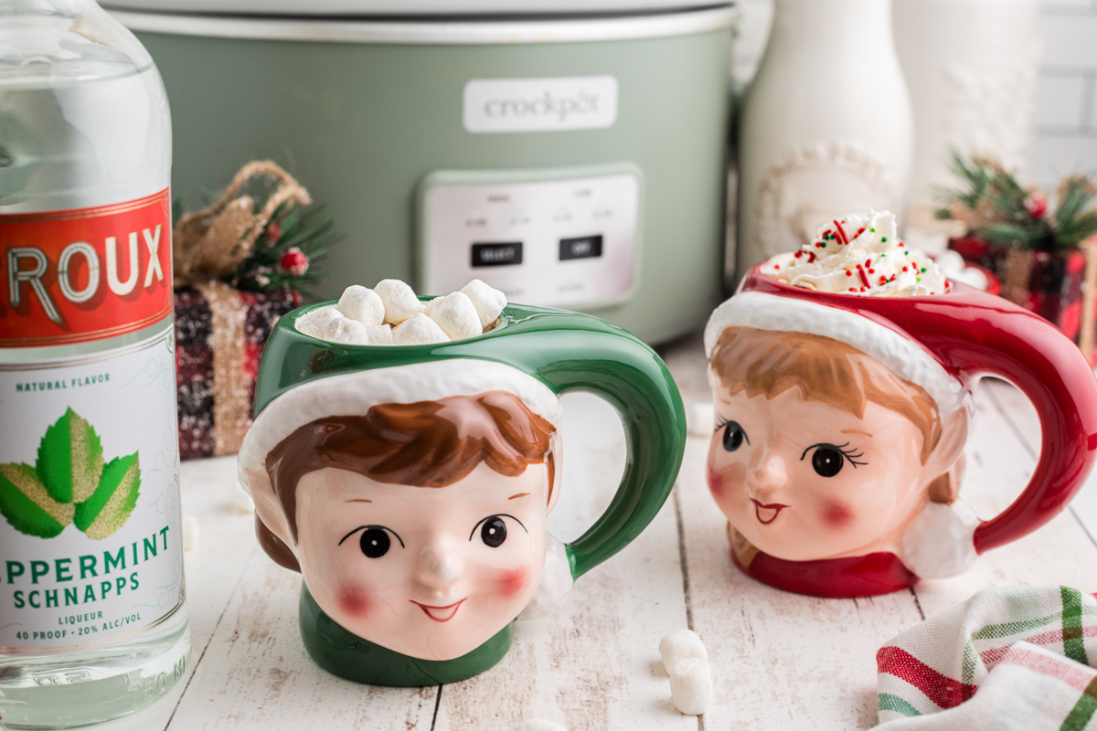 Slow cooker boozy hot chocolate in festive mugs in front of a crockpot.