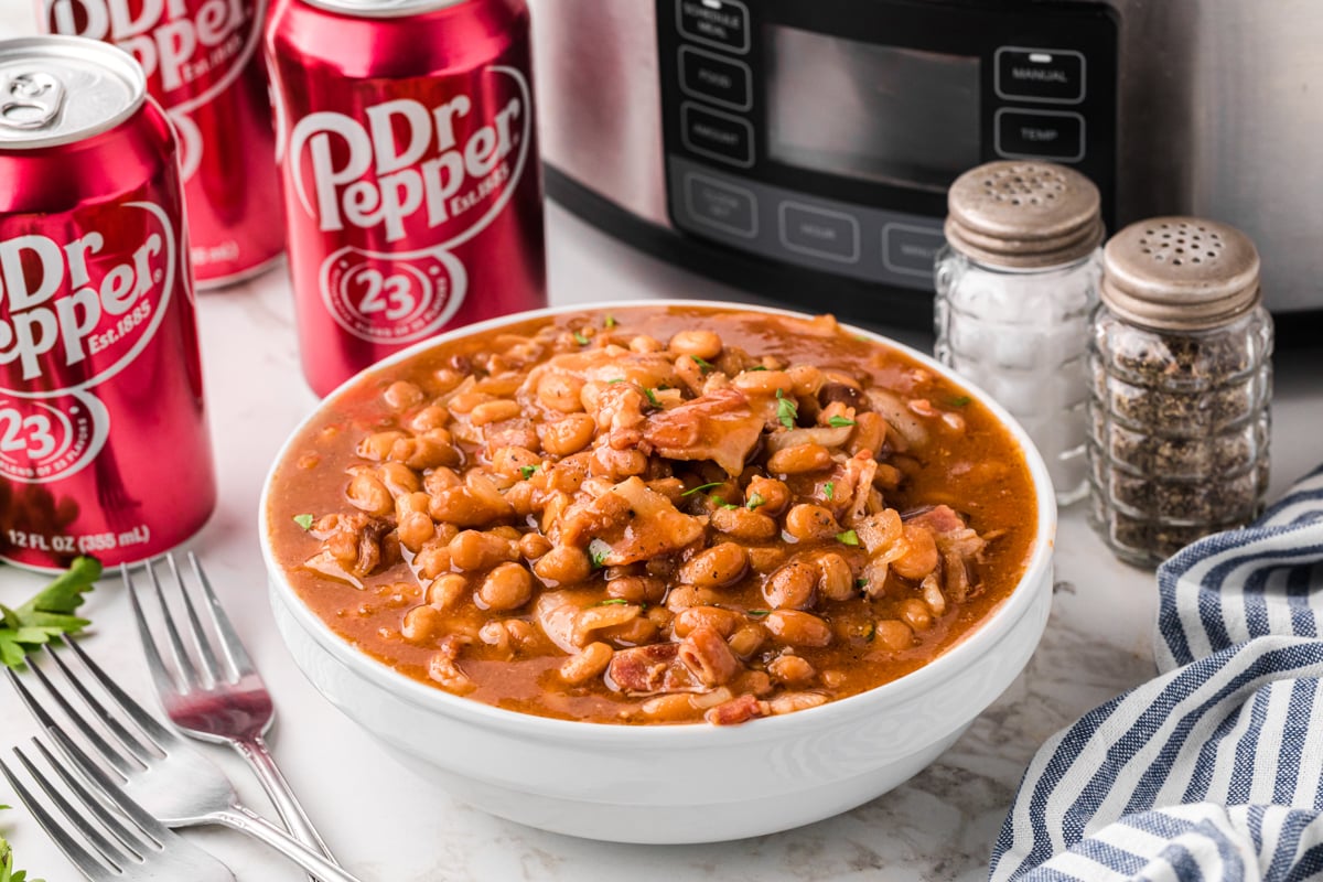 slow cooker dr. pepper baked beans in a white bowl with cans of dr. pepper in the background.