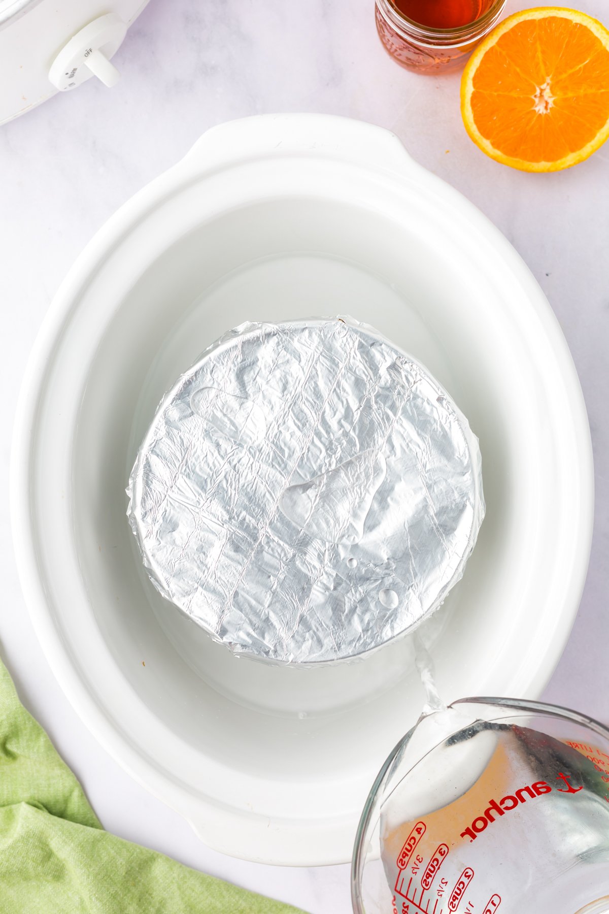 Foil-covered figgy pudding in a crockpot with water being poured around it.