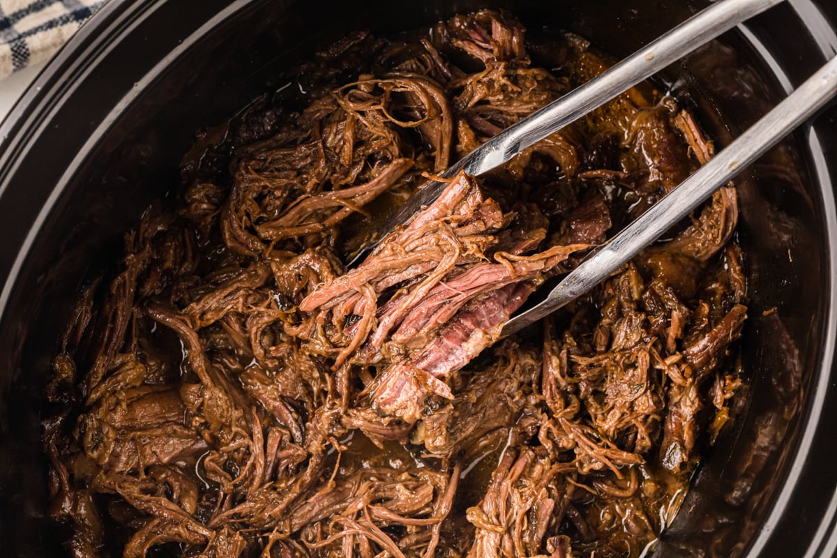 A pair of tongs pulling some slow cooker georgia pot roast out of a crockpot.