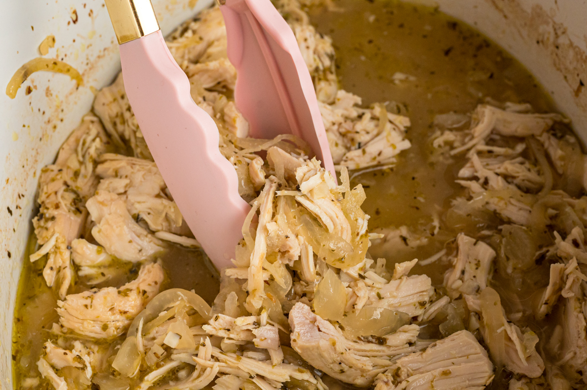 A pair of tongs grabbing some meat for slow cooker chicken gyros.