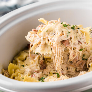 A scoop of slow cooker chicken cordon bleu pasta being held up over a crockpot.