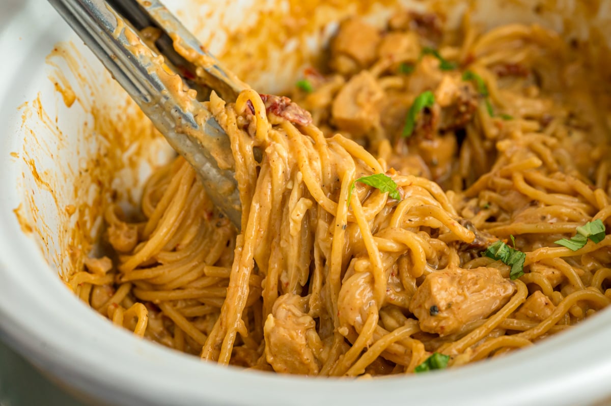 Tongs grabbing slow cooker marry me chicken pasta from a crockpot.
