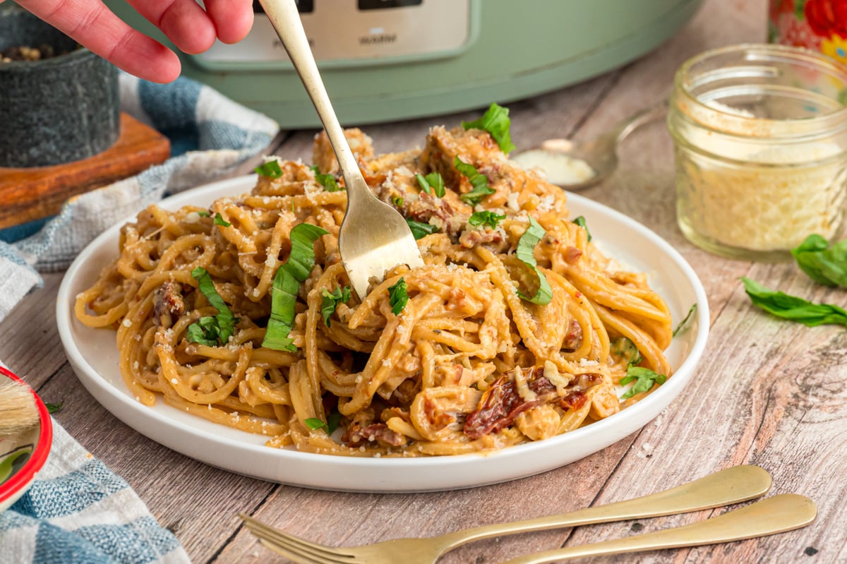 A hand holding a fork grabbing some slow cooker marry me chicken pasta from a plate.