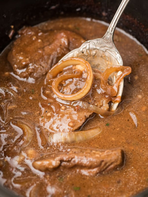A large spoon scooping out steak and onions from a slow cooker.