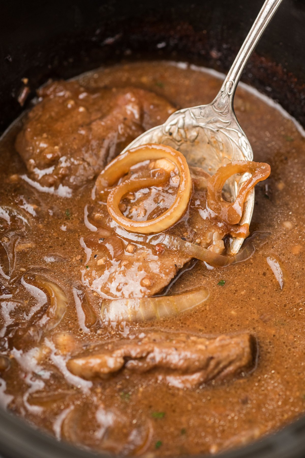 A large spoon scooping out steak and onions from a slow cooker.