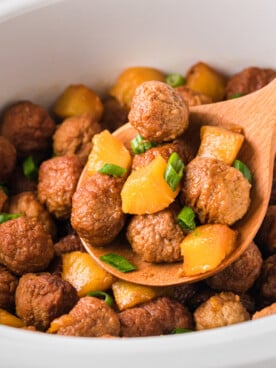 A wooden spoon holding a scoop of pineapple meatballs over a crockpot.