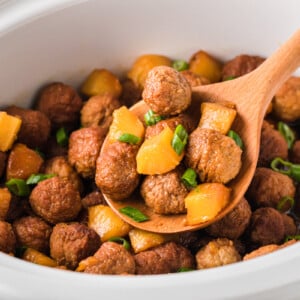 A wooden spoon holding a scoop of pineapple meatballs over a crockpot.