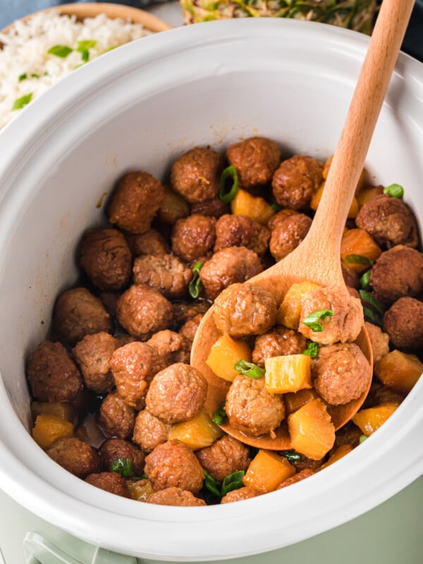 A crockpot full of pineapple meatballs with a wooden spoon scooping some out.