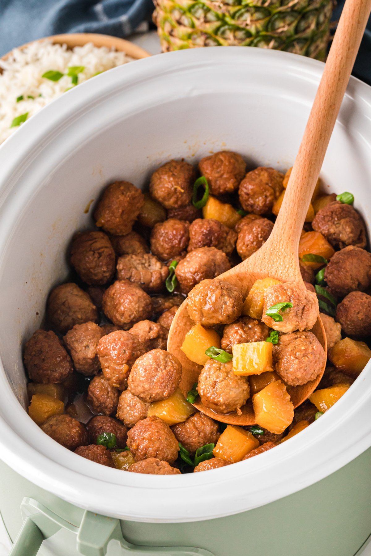 A crockpot full of pineapple meatballs with a wooden spoon scooping some out.
