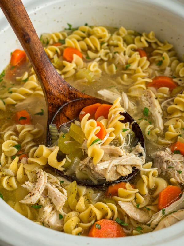 Slow cooker chicken noodle soup with a wooden spoon in it.
