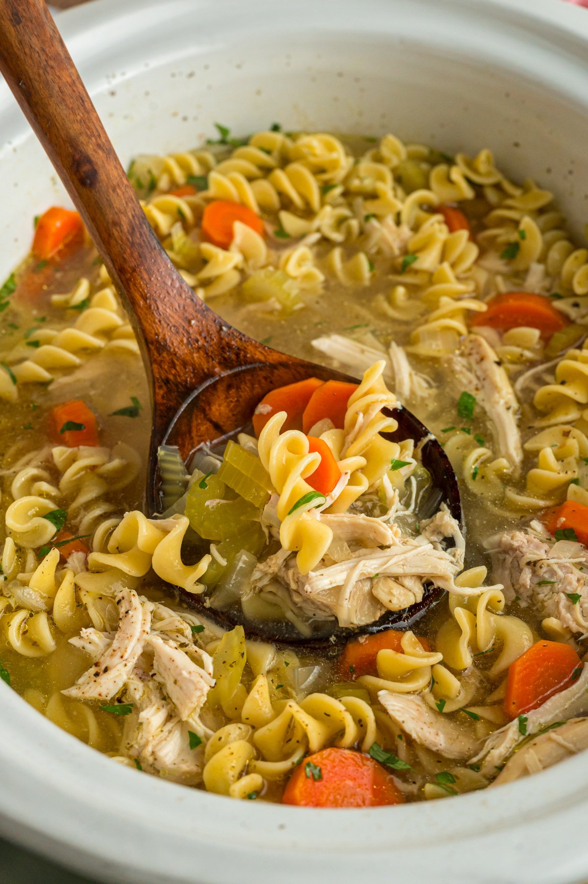 Slow cooker chicken noodle soup with a wooden spoon in it.