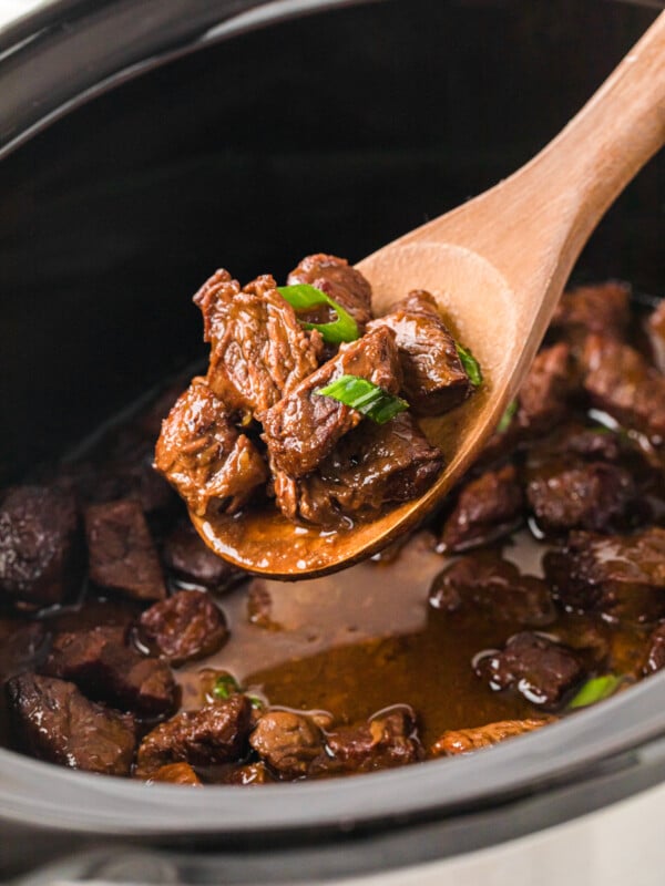 A wooden spoon scooping honey garlic steak bites out of a crockpot.