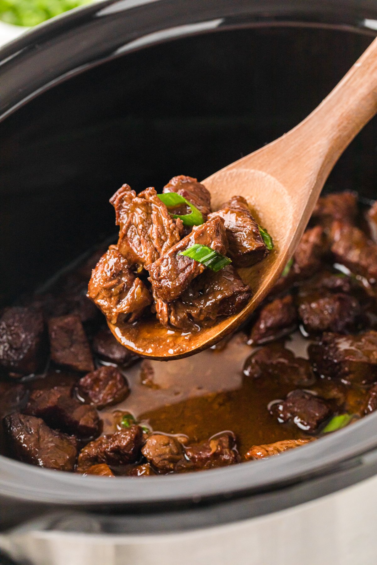 A wooden spoon scooping honey garlic steak bites out of a crockpot.