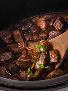 Cooked steak bites in a slow cooker with a wooden spoon in it.