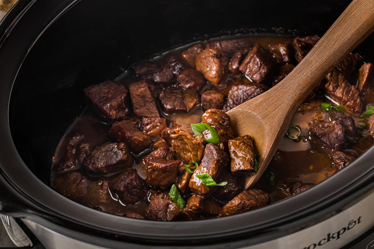 Cooked steak bites in a slow cooker with a wooden spoon in it.