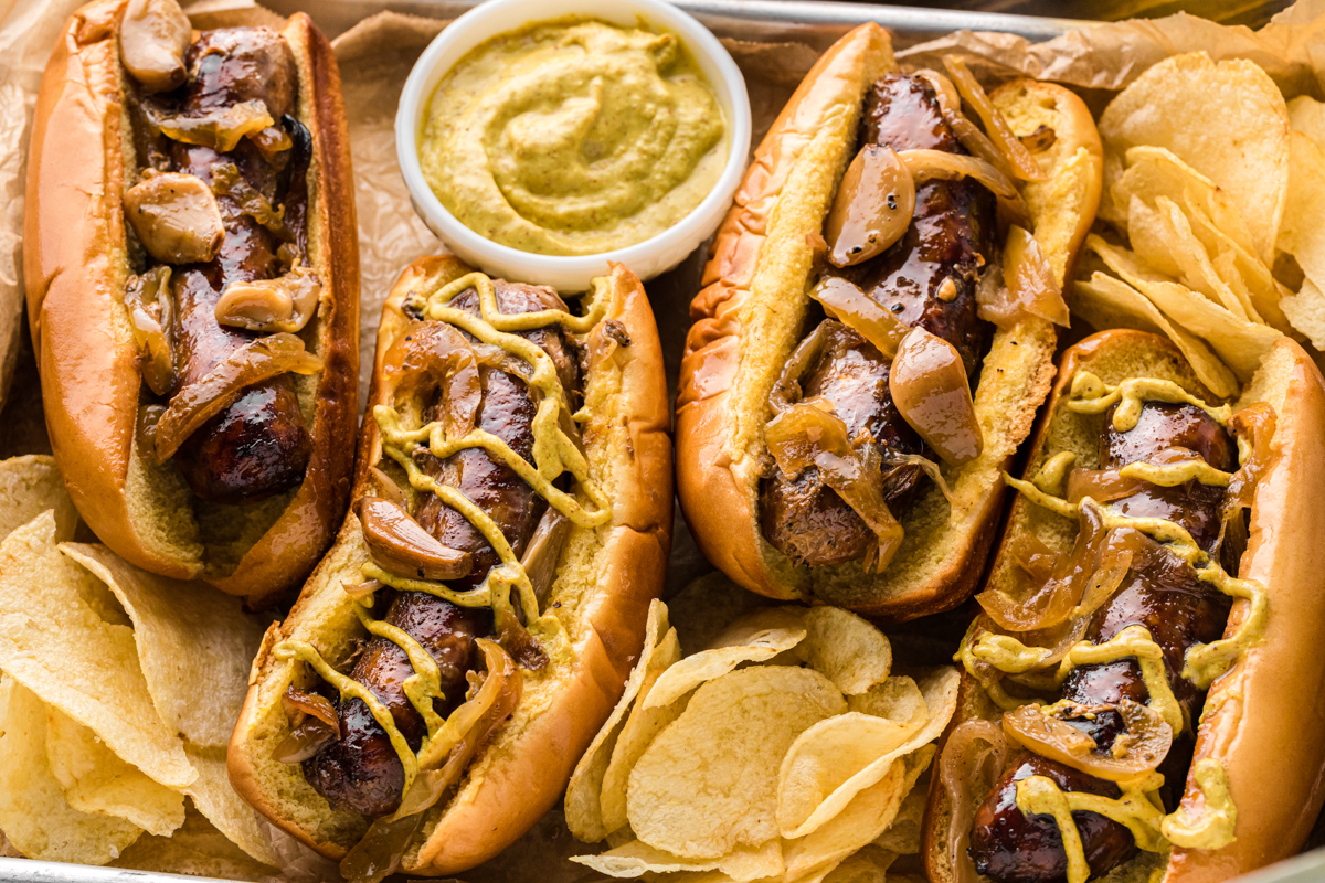 Close-up of brats in buns with mustard and potato chips around them.