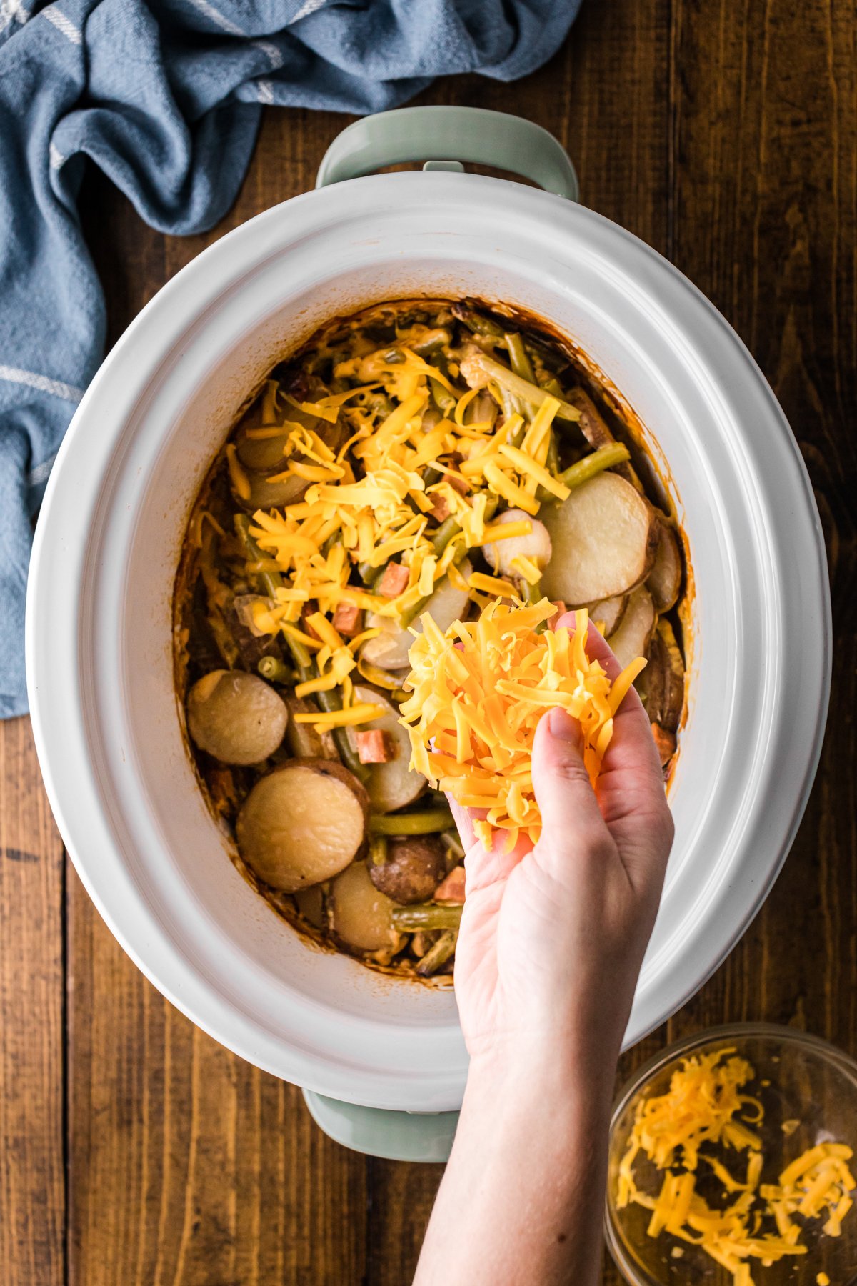 Shredded cheese being added to ham and potato dinner in a crockpot.