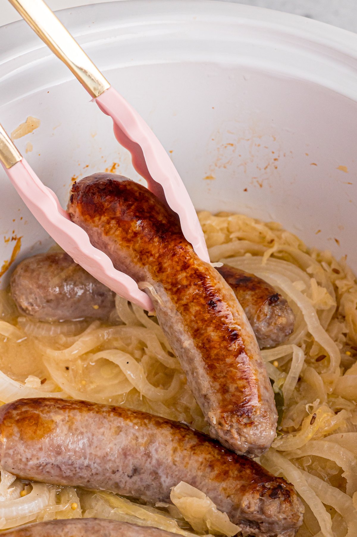 Tongs picking up a bratwurst from a crockpot with sauerkraut in it.